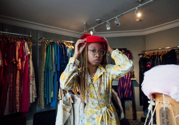 A young woman enjoying a day out shopping in a vintage clothing store in Durham, North East England. She is standing in the shop, trying on a red wedding hat while looking at the camera and posing.