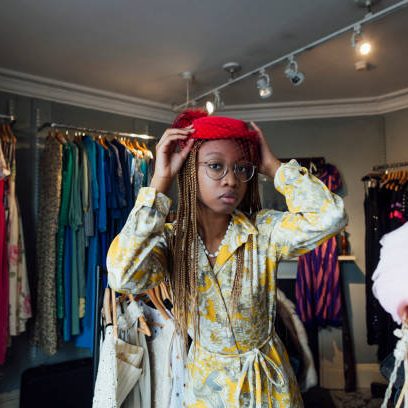 A young woman enjoying a day out shopping in a vintage clothing store in Durham, North East England. She is standing in the shop, trying on a red wedding hat while looking at the camera and posing.
